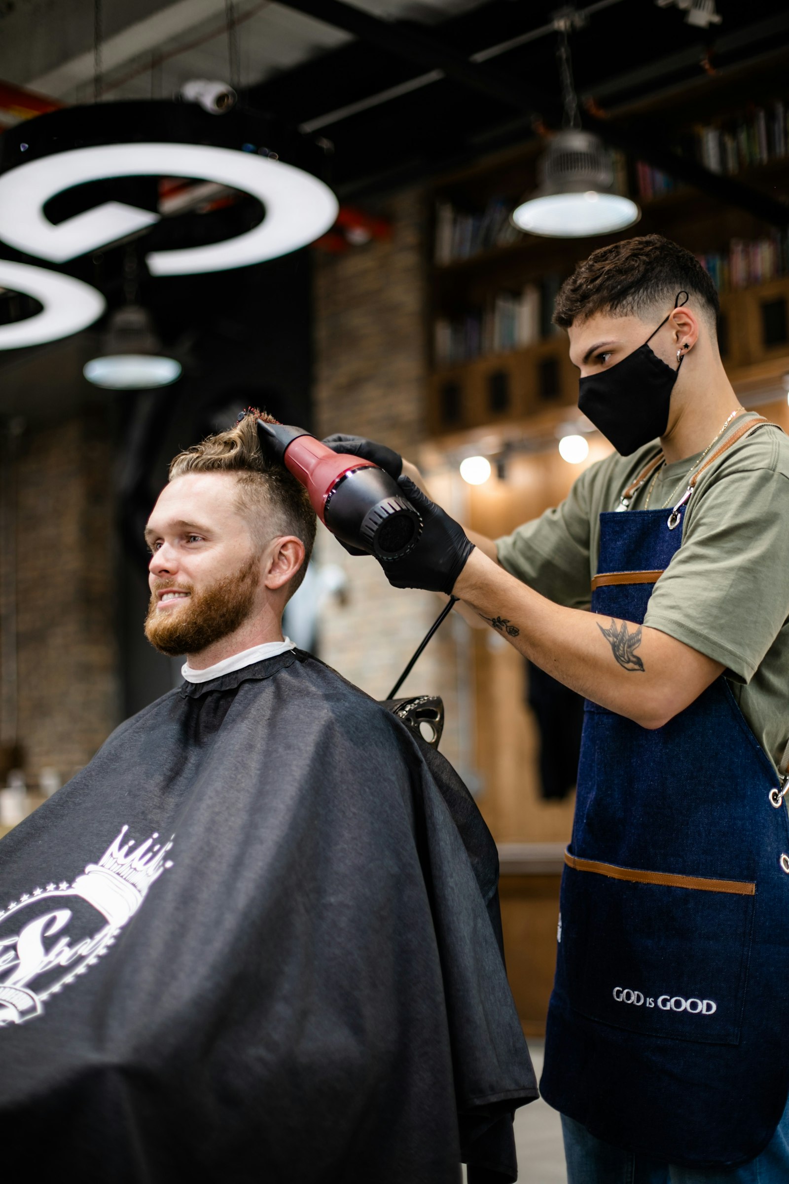 A barber in a denim apron blow-drying a client's hair